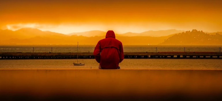 A depressed man sitting by a body of water in the evening.