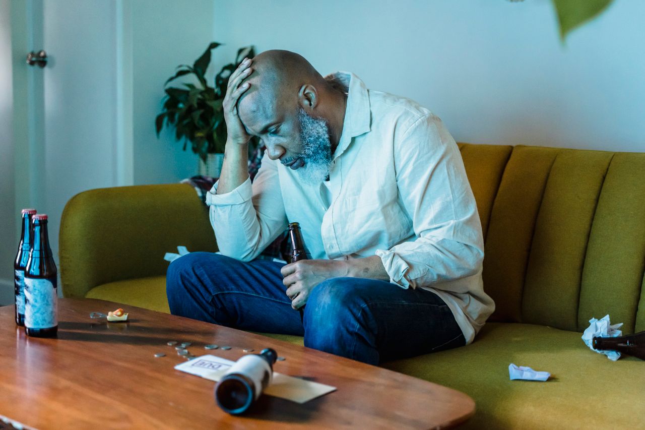 Troubled man sitting on a sofa in a cluttered living room with his head resting in his hand.
