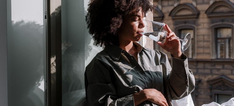 Pregnant woman drinking a glass of water while sitting by a window.