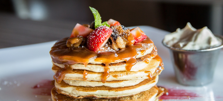 A tower of pancakes with caramel, a side of whipped cream, blueberries, and strawberries on top.