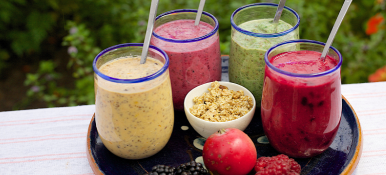 Four smoothies of different colors, a small plate of granola in the middle, an apple, and raspberries.