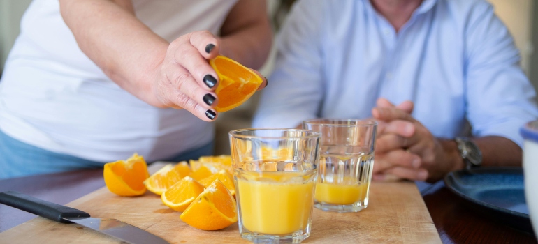 A woman is squeezing an orange in a glass and talking to a man about nutrition and addiction recovery.