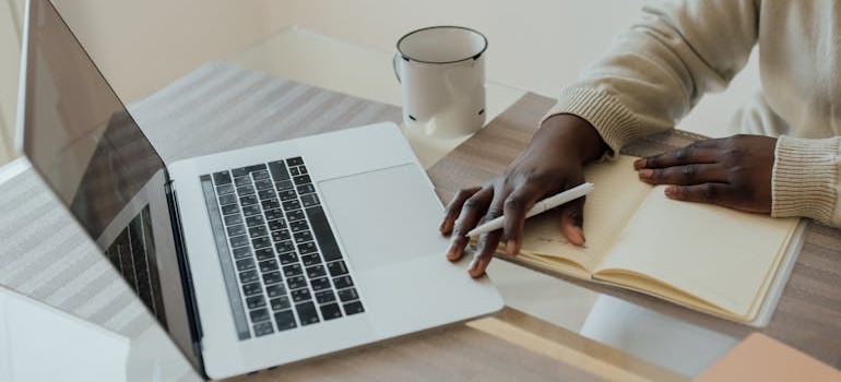 Man writing in a notebook and looking at his laptop.