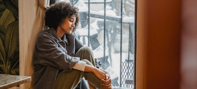 Woman sitting on the windowsill and experiencing holiday blues symptoms.