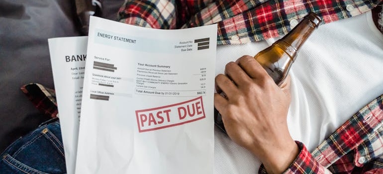 Man holding a bottle of beer with unpaid bills next to him.