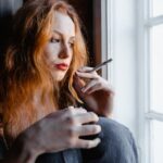 Woman looking out of a window and smoking a cigarette while struggling with depression and addiction.
