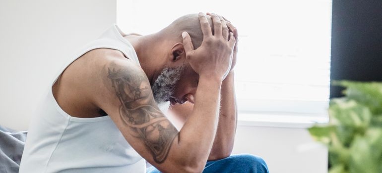 Man sitting on a bed holding his head while experiencing physical distress.