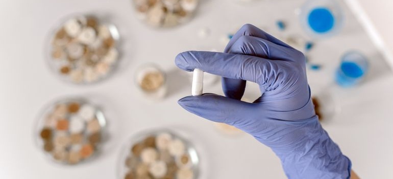 Close-up of a blue-gloved hand holding a white capsule in a laboratory.