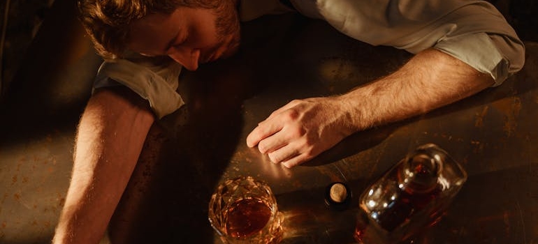 Man falling asleep on the table with a glass and bottle of whiskey next to him.