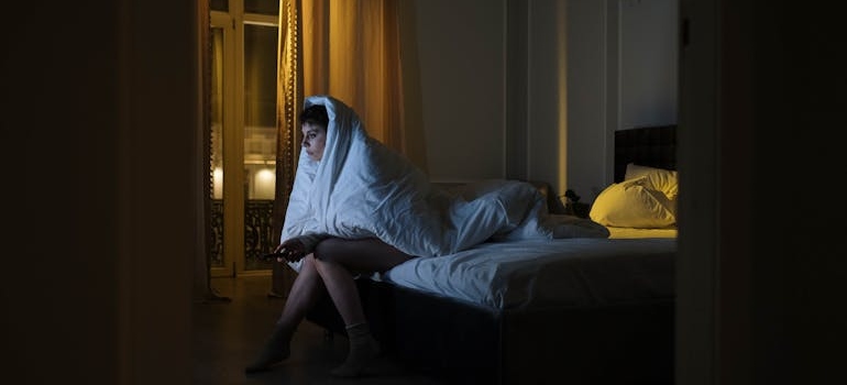 Woman struggling with insomnia while sitting on the bed and covering herself with a white blanket.