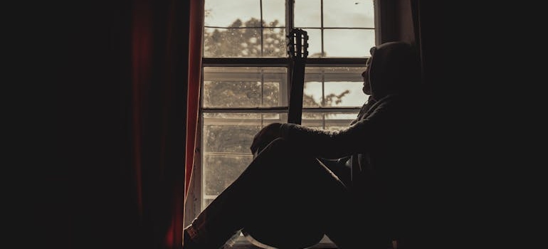 Man holding a guitar and sitting near a window.