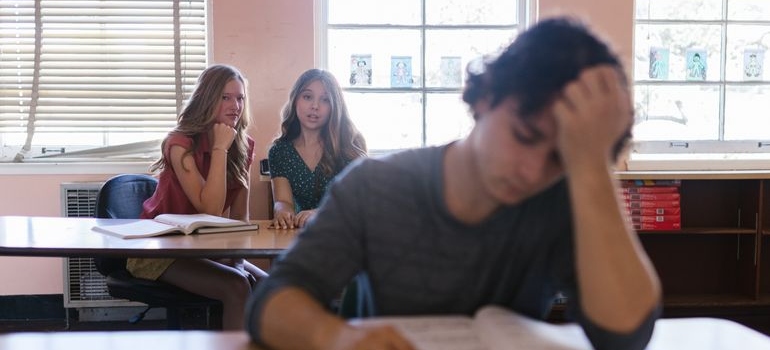 Two girls gossiping about a boy sitting in front of them.